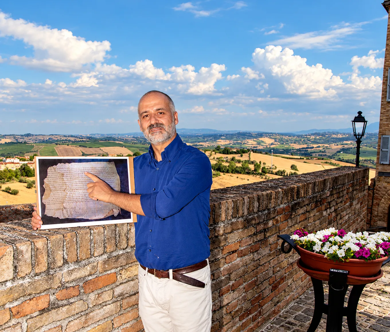 Iuri Silvestri on a brick terrace overlooking the hills, pointing to a photograph of a parchment.